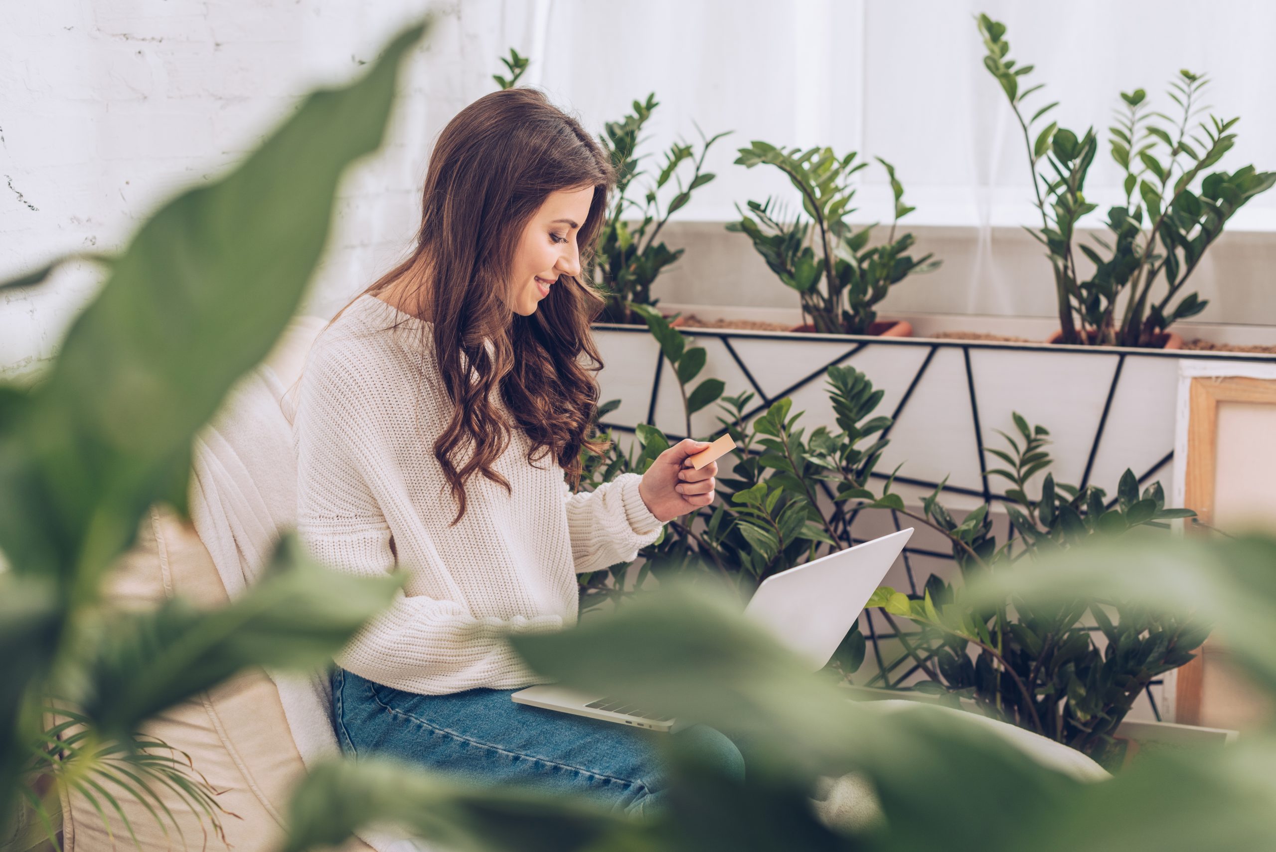 smiling woman holding credit card and looking at laptop siting among houseplants